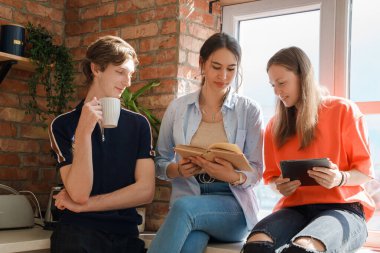 Portrait of group of three friends reading book and using tablet around window at home.