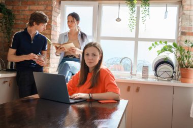Portrait of woman using laptop at table and her two friends reading book at her back.