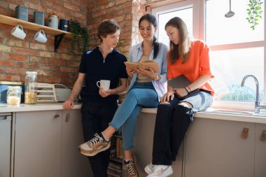 Portrait of group of three friends reading book and using tablet around window at home.