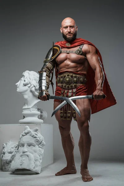 Portrait of greek gladiator from past holding swords posing around greek busts.