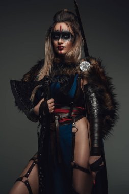 Portrait of female warrior dressed in attire and fur holding axe against grey background.