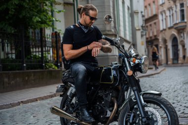 Dövmeli havalı bir motorcunun, sokaklarda bisiklet sürerken saatlere baktığı bir fotoğraf..