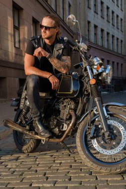 Gün batımında motorsikletinin üstünde rockçı gibi poz veren şık bir motosikletlinin fotoğrafı..