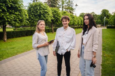 Yazın şehir parkında dizüstü bilgisayar ve tabletle gezen üç öğrencinin fotoğrafı..