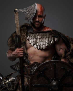 Yüzü kanlı, kaslı viking vuruşu. Gri arka planda balta ve kalkan tutuyor..