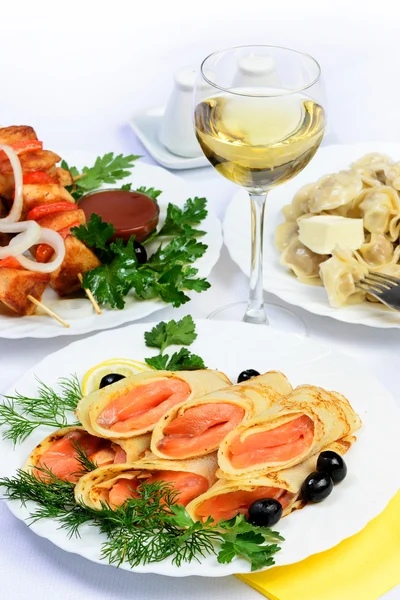Table with food of meat, salmon rolls, dumplings and white wine. — Stock Photo, Image
