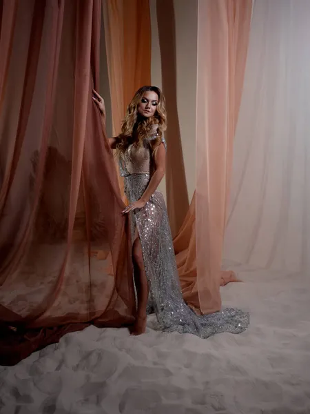 Una joven rubia elegante con un vestido translúcido brillante detrás de una cortina —  Fotos de Stock
