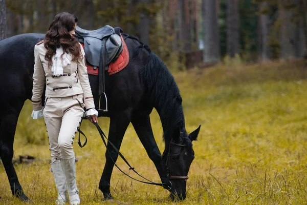 Una joven morena hermosa jinete junto a un caballo negro en plena munición, —  Fotos de Stock