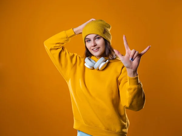 Stylowa młoda brunetka w żółtej bluzie z kapturem i kapeluszach pokazuje fajny gest, — Zdjęcie stockowe