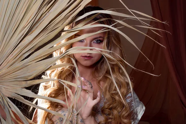 Eine schöne junge Frau mit hellem Make-up versteckt sich im Schatten eines Palmenzweiges. — Stockfoto