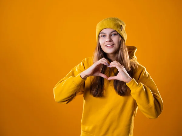 Elegante alegre jovem morena em um capuz amarelo e chapéus mostra um sinal de coração — Fotografia de Stock