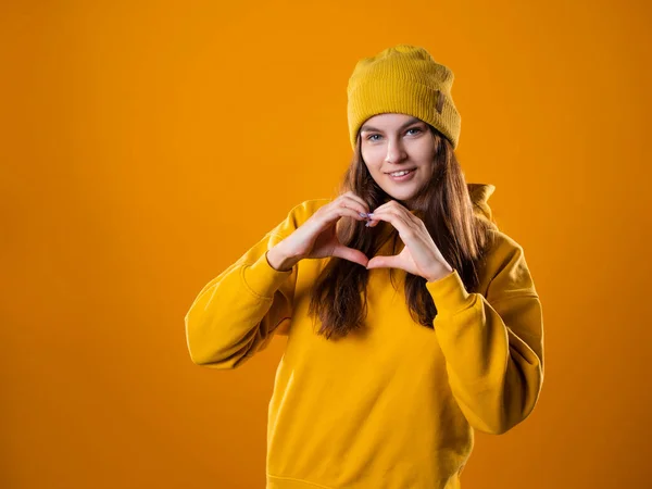 Stylová veselá mladá brunetka ve žluté mikině a klobouky ukazuje srdeční znamení — Stock fotografie