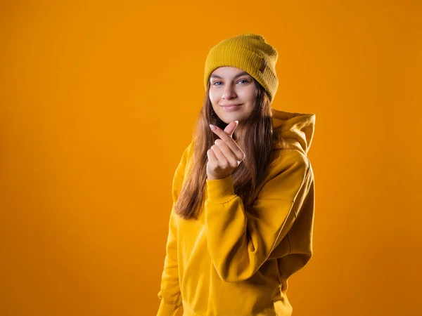 Stijlvolle vrolijke jonge brunette in een gele hoodie en hoeden toont een hart teken — Stockfoto