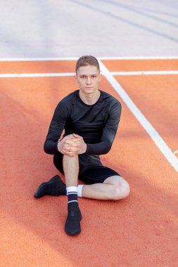 Genç, yakışıklı bir adam spor sahasında oturup kameraya bakıyor.