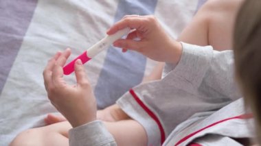 Mutlu kadın yatakta yatıyor ve hamilelik testi yapıyor. Tanımlanamayan bir kız iki çizgili pozitif hamilelik testi tutuyor. Hamilelik konsepti, aile planlaması.