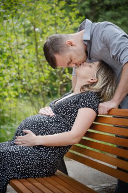 Kocası parktaki bankta oturan hamile bir kadın. Adam karısını öpüyor. Sevgili çift birlikte eğleniyor - bebeğin doğmasını bekliyor.