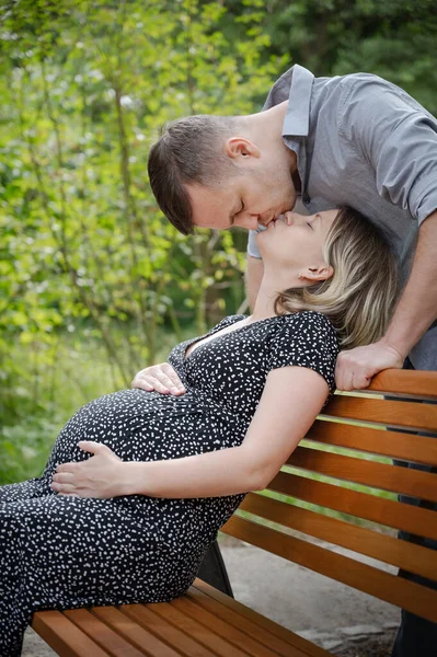 Kocası parktaki bankta oturan hamile bir kadın. Adam karısını öpüyor. Sevgili çift birlikte eğleniyor - bebeğin doğmasını bekliyor.