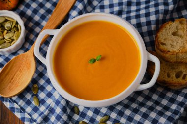 Mavi kareli peçete, kızarmış ekmek, kabak tohumu ve kırsal ahşap arka planda fesleğen üzerine beyaz bir bulyon içinde ev yapımı balkabağı kreması çorbası seçici bir odak noktası.