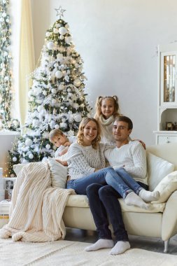 Güzel, mutlu bir ailenin portresi. Beyaz süveterli iki çocuk kanepeye oturmuş Noel ağacıyla süslenmiş bir odada.