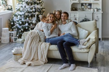 Güzel, mutlu bir ailenin portresi. Beyaz süveterli iki çocuk kanepeye oturmuş Noel ağacıyla süslenmiş bir odada.