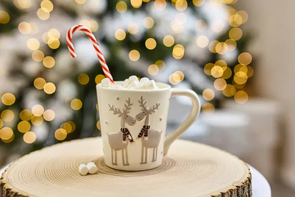 Fairy lights background, hot cocoa with marshmallows, white cup with deer on a wooden cut at home during Christmas and New Year holidays. Soft focus bokeh.