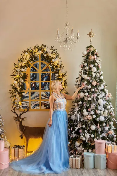 Mavi uzun elbiseli güzel sarışın kadın parlak bir odada duruyor. Parlak Noel ağacını süslüyor. İçi geyik oyuncakları, çelenkler, fenerler, mumlar, hediyelerle dolu.