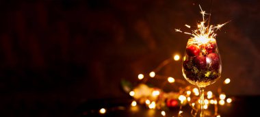 Şarap kadehinde yanan bengal havai fişekleri ve arka planda kırmızı Noel topları. Yaratıcı estetik an. Yeni yıl atmosferi ve gece havası. Seçici odaklanma. Pankart