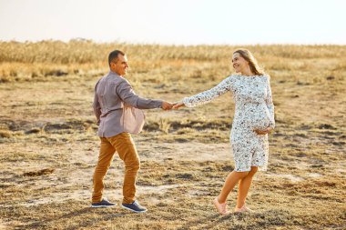 Koca göbekli hamile koca güneşli yaz mevsiminde nehir kıyısında dinleniyor. Mutlu çift el ele tutuşmaya bayılıyor. Beyaz erkek ve kadın dışarıda el ele tutuşuyorlar.