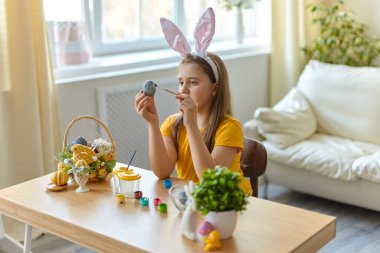 Tavşan kulağı takan şirin bir çocuk. Oturma odasındaki masada oturan kız. Çocuk yumurta boyuyor..