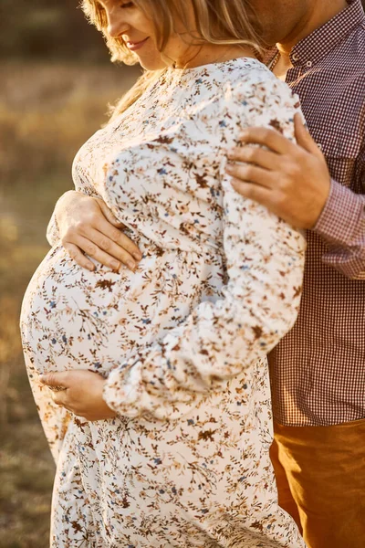 未来母亲的手紧紧抓住怀孕的大肚子 丈夫拥抱怀孕的妻子 夏日日落时 欢乐的家人在大自然中依偎着 身穿白色棉衣的白人妇女 — 图库照片