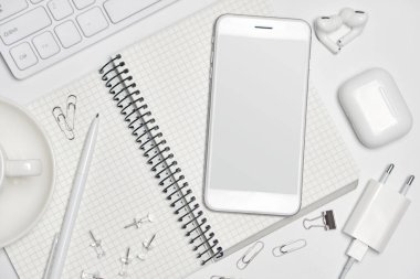 Ofis masasının çalışma alanının en üst görüntüsü. Akıllı telefon boş ekran ve boş not defteri, kahve fincanı, kulaklık, şarj aleti, klavye beyaz arka planda fotokopi alanı. Yaratıcı Tasarımcı kavramı.