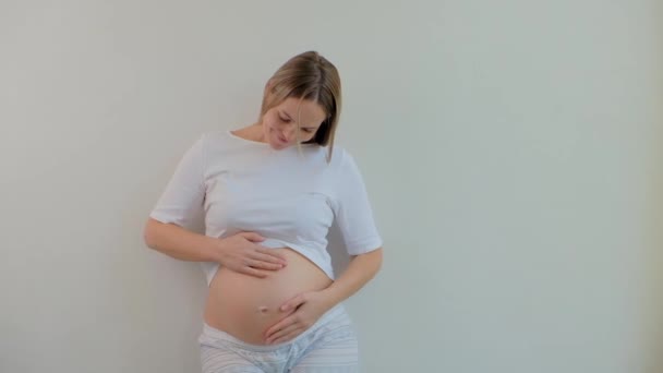 Όμορφη Έγκυος Γυναίκα Πιτζάμες Στέκεται Στο Φόντο Ενός Λευκού Τοίχου — Αρχείο Βίντεο