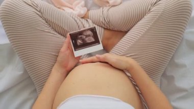 Hamile bir kadının evde bebek kıyafetleri hazırlayıp ultrason fotoğrafına, hamilelik ve doğum konseptine baktığı en iyi manzara.
