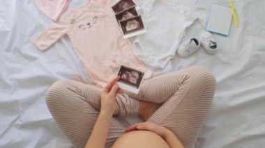 Hamile bir kadının evde bebek kıyafetleri hazırlayıp ultrason fotoğrafına, hamilelik ve doğum konseptine baktığı en iyi manzara.