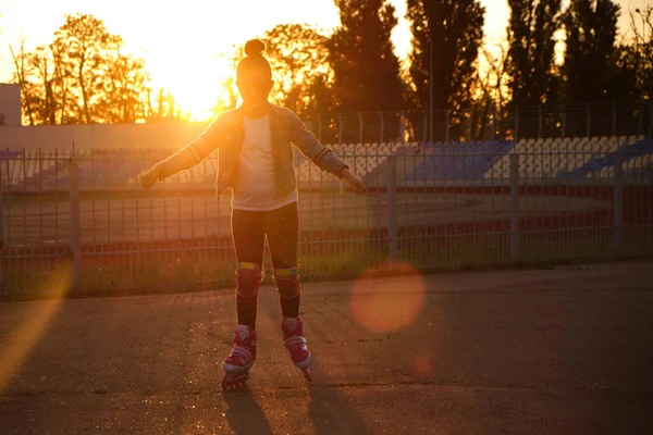 Little pretty happy funny girl on roller skates at stadium at sunset, learning to roller skate outdoors. Outdoor activity for children. Active sport for preschool kid. sun glare, selective focus