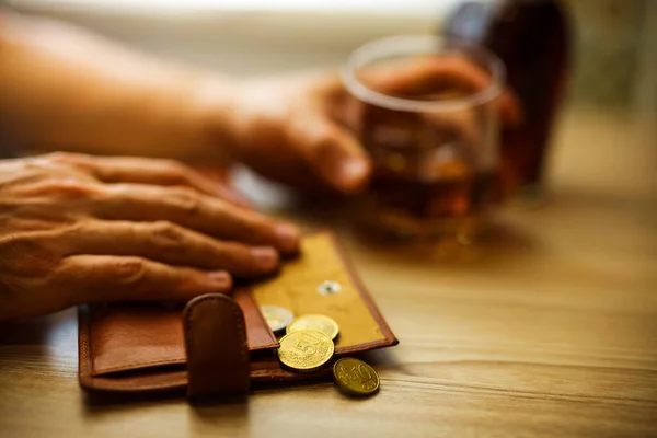 Fired man countsing his last money to drink expensive alcohol. Wrinkled alcoholic sits at brown wooden table with a glass of cognac with ice, on the table, empty wallet with coins