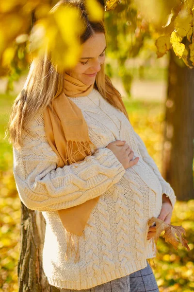 Attractive young pregnant woman with leaf holding her tummy in autumn park. Beautiful future mother having great time outside on sunny fall day and enjoying beautiful nature outdoors