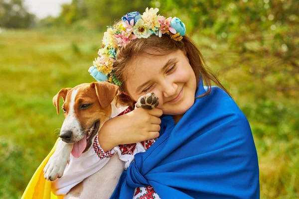 Ukrainian little girl in national clothes, covered with a blue-yellow flag, hugs her dog, dressed in national Ukrainian vyshyvanka. The concept of patriotism and national identity. Support Ukraine