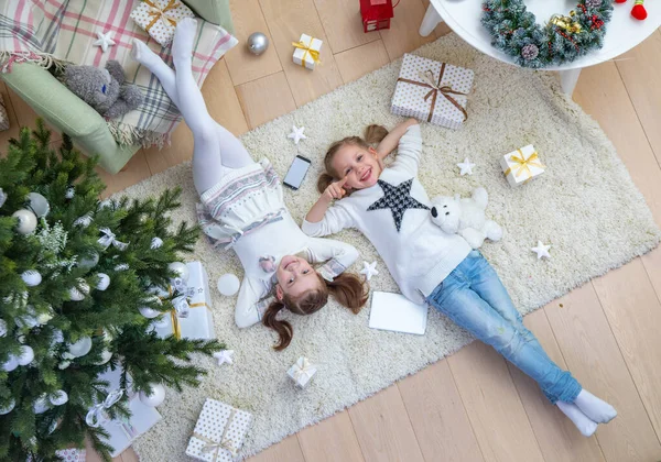 2人の幸せな少女がクリスマスツリーの下に横たわってカーペットの上で贈り物をしています上からの眺め — ストック写真