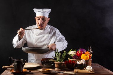 Beyaz üniformalı erkek aşçı, lokantanın mutfağında servis yapmadan önce ahşap kepçeyle yemek tadıyor.