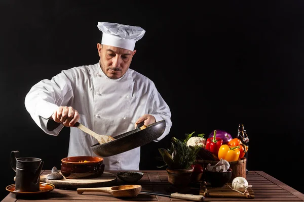 Male cook in white uniform and hat with ladle mixes the ingredients onto the cooking pan before serving while working in a restaurant kitchen