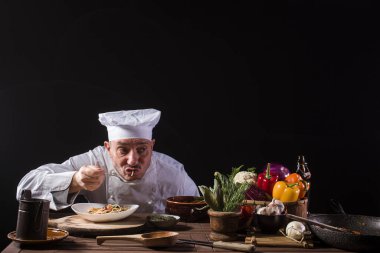 Restoran mutfağında beyaz üniformalı bir erkek şef servis etmeden önce taze sebzeli spagetti tadıyor.