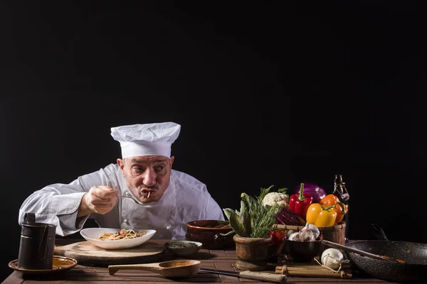 Male chef in a restaurant kitchen wearing white uniform tasting spaghetti with fresh vegetables before serving it