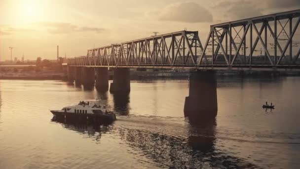 Motorboot-Yacht fahren Fluss. Eisenbahnbrücke im Hintergrund. Leuchtend orangefarbener Sonnenuntergang über Segelboot — Stockvideo