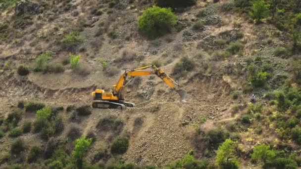 Világos sárga kotró ássa a sziklát a hegygerincen. Útépítés dombokon, sárga homokos földön. — Stock videók