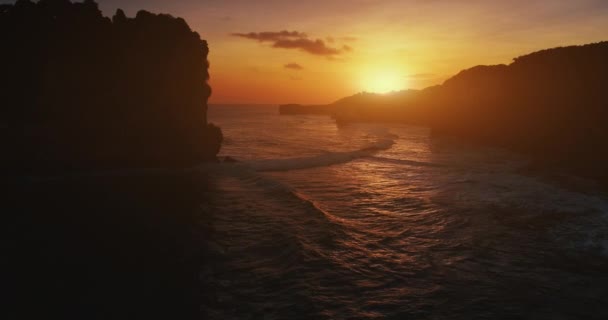 オレンジの夕日の崖の海岸シルエット オーシャンベイの波はゆっくりとした動きの空中ビュー 海の水の背景に岩の山を閉じます 黄色の夜の太陽の下で熱帯岩の海岸 夏の旅行バリ インドネシア — ストック動画