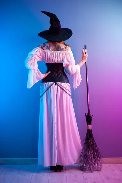 Una chica en un disfraz de bruja para Halloween con una escoba y una calabaza en luz de neón —  Fotos de Stock