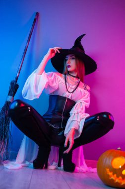 Cadılar Bayramı için cadı kostümü giymiş bir kız elinde süpürge ve neon ışıklı bir balkabağıyla.
