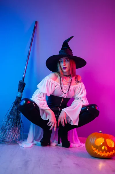 Een meisje in een heks kostuum voor Halloween met een bezem en een pompoen in neon licht — Stockfoto