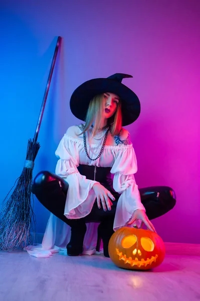 Una chica en un disfraz de bruja para Halloween con una escoba y una calabaza en luz de neón —  Fotos de Stock